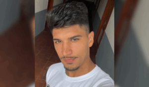 Homem jovem com cabelo escuro e curto faz selfie em um corredor com piso de cerâmica vermelha.