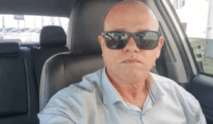 Homem calvo de óculos escuros tirando uma selfie dentro de um carro, usando camisa social clara.