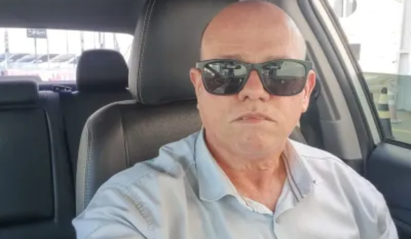 Homem calvo de óculos escuros tirando uma selfie dentro de um carro, usando camisa social clara.