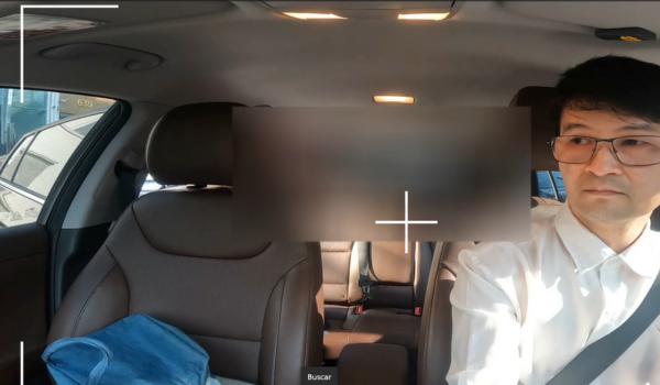 Motorista de aplicativo de camisa branca e óculos dirige durante o dia com o carro vazio, registrado por uma câmera interna no painel.