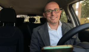 Homem sorridente de óculos, vestindo blazer cinza e camisa branca, sentado no banco do motorista de um carro, com um celular posicionado à frente do volante.