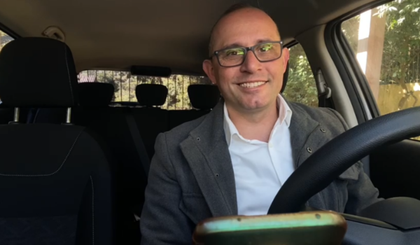 Homem sorridente de óculos, vestindo blazer cinza e camisa branca, sentado no banco do motorista de um carro, com um celular posicionado à frente do volante.