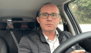 Homem de óculos e jaqueta preta, sentado ao volante dentro de um carro, olhando diretamente para a câmera.