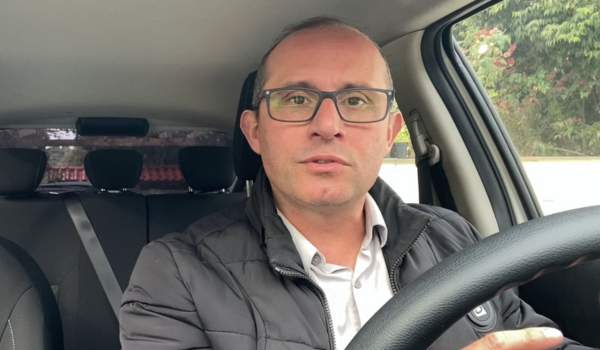 Homem de óculos e jaqueta preta, sentado ao volante dentro de um carro, olhando diretamente para a câmera.