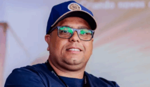 Homem sorridente de óculos e boné azul, com camiseta escura e crachá, em ambiente interno com fundo desfocado.