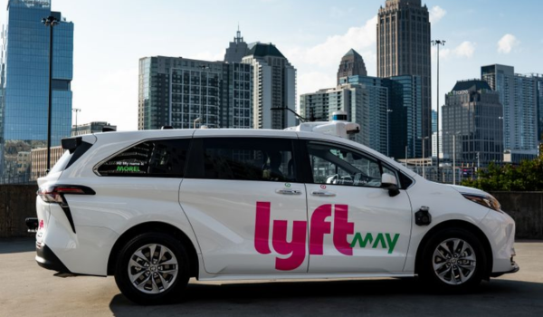 Carro autônomo branco da Lyft operado pela empresa May Mobility, com logotipos nas portas, estacionado em área urbana com arranha-céus ao fundo.