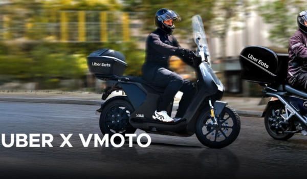 Dois entregadores usando scooters elétricas com baú do Uber Eats em uma rua urbana, com a legenda "Uber x Vmoto".