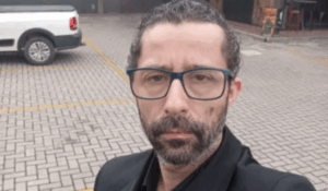 Homem de barba curta, óculos de armação azul escura e roupa social preta, posando sério em estacionamento de piso intertravado, com uma caminhonete branca ao fundo.