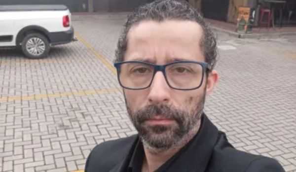 Homem de barba curta, óculos de armação azul escura e roupa social preta, posando sério em estacionamento de piso intertravado, com uma caminhonete branca ao fundo.