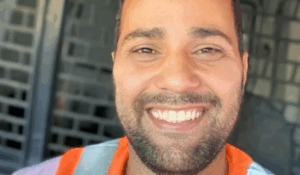 Homem sorridente vestindo uniforme com detalhes em laranja e lilás, posando para uma selfie em ambiente interno.