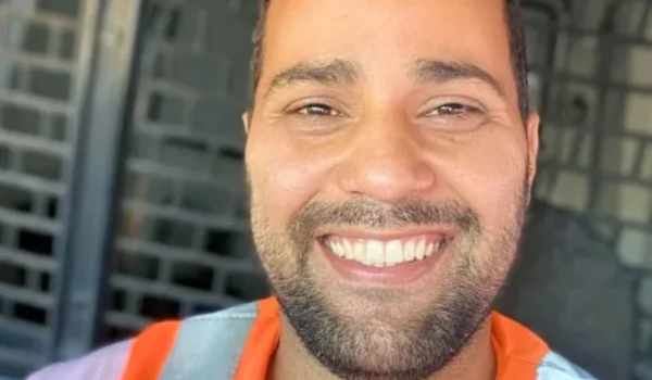 Homem sorridente vestindo uniforme com detalhes em laranja e lilás, posando para uma selfie em ambiente interno.