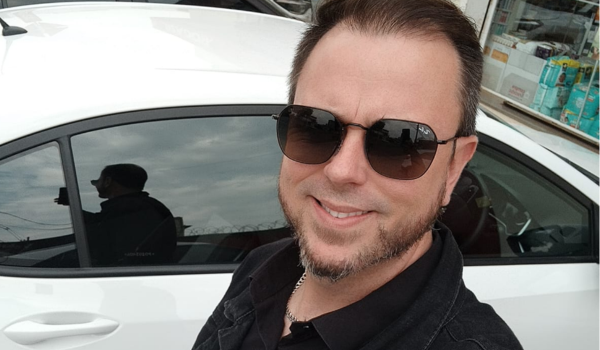 Homem branco sorrindo, usando óculos escuros e camisa preta, posando em frente a um carro branco.