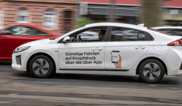 Carro branco com publicidade da Uber em alemão circulando por uma rua urbana, com motorista visível ao volante.