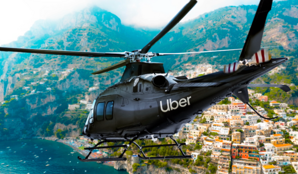 Helicóptero preto com a marca Uber sobrevoando uma cidade costeira com construções em encosta e mar ao fundo.