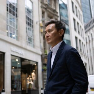 Homem de meia-idade vestindo blazer azul-marinho e camisa branca, caminhando por uma rua com prédios comerciais de arquitetura clássica e moderna.