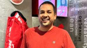 Homem sorridente usando camiseta vermelha em um ambiente moderno com palavras motivacionais na parede ao fundo.