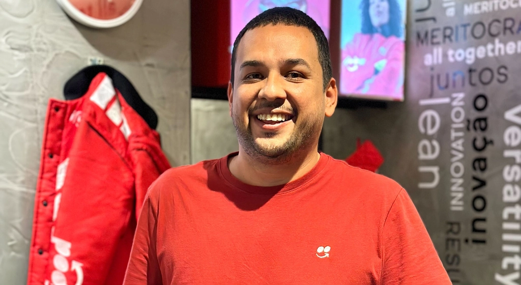 Homem sorridente usando camiseta vermelha em um ambiente moderno com palavras motivacionais na parede ao fundo.