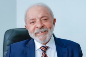 Homem idoso de barba branca, vestindo terno azul e gravata listrada, sorrindo para a câmera em um ambiente interno.