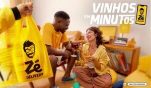 Pessoa entregando uma sacola amarela do Zé Delivery para um casal sorridente, que brinda com taças de vinho em uma sala com decoração amarela.