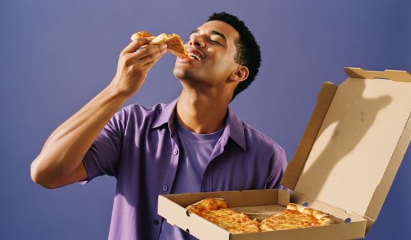 Homem sorrindo enquanto come uma fatia de pizza, segurando uma caixa de pizza aberta.
