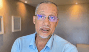 Homem de pele parda e cabelos curtos grisalhos, usando óculos com lentes parcialmente reflexivas e camisa azul clara, olhando diretamente para a câmera em um ambiente interno com iluminação suave.