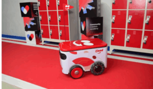 Robô de entregas do iFood em corredor com armários vermelhos, ao lado de um painel da Community Creators Academy.
