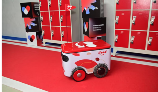 Robô de entregas do iFood em corredor com armários vermelhos, ao lado de um painel da Community Creators Academy.