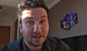 Homem com barba e cabelo escuro, olhando para a câmera em um ambiente interno iluminado pelo sol, com um pôster colorido dos personagens Rick e Morty na parede ao fundo.