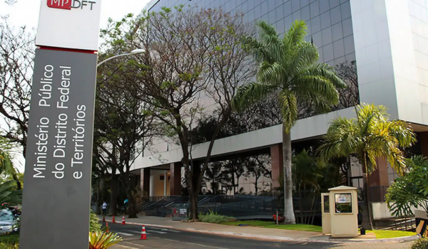 Fachada do edifício do Ministério Público do Distrito Federal e Territórios (MPDFT), com uma placa institucional na entrada e árvores ao redor do prédio envidraçado.