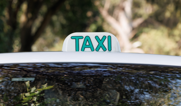 Placa luminosa com a palavra "TÁXI" em letras verdes, fixada no teto de um carro branco.