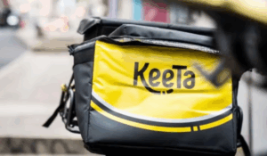 Bolsa térmica de entregas da empresa Keeta com design em amarelo e preto sobre uma motocicleta em uma calçada urbana.