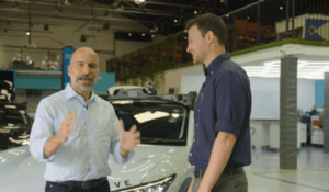 Dois homens conversam em pé na frente de um carro autônomo branco em ambiente de laboratório tecnológico.