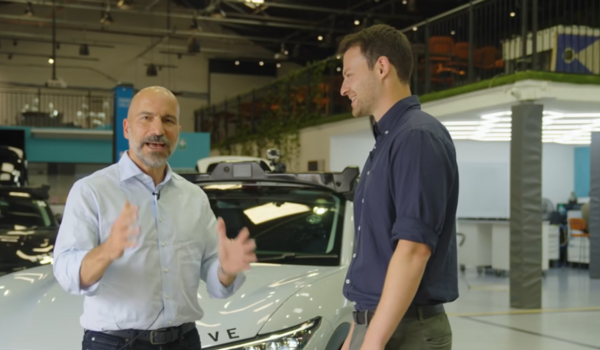 Dois homens conversam em pé na frente de um carro autônomo branco em ambiente de laboratório tecnológico.