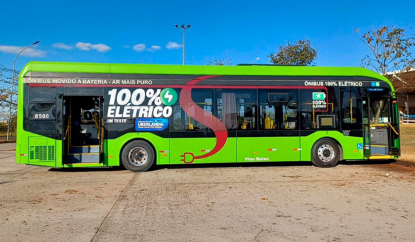 Ônibus verde de transporte público, com a mensagem "100% Elétrico" estampada na lateral, indicando que é movido à bateria. O veículo está parado em uma área aberta sob céu azul.