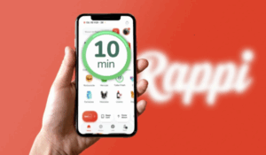 Pessoa segura um smartphone exibindo o aplicativo da Rappi, com destaque para um selo verde indicando entrega em “10 min”. Ao fundo, o logotipo da Rappi desfocado sobre um fundo vermelho.