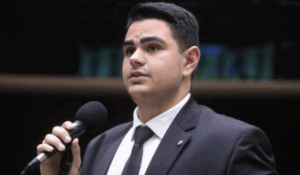 Homem jovem de terno e gravata fala ao microfone em ambiente formal, como uma tribuna parlamentar.