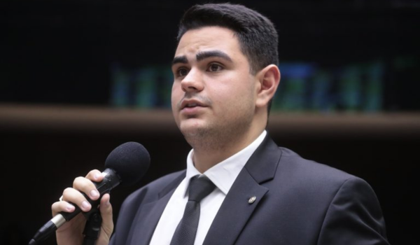 Homem jovem de terno e gravata fala ao microfone em ambiente formal, como uma tribuna parlamentar.