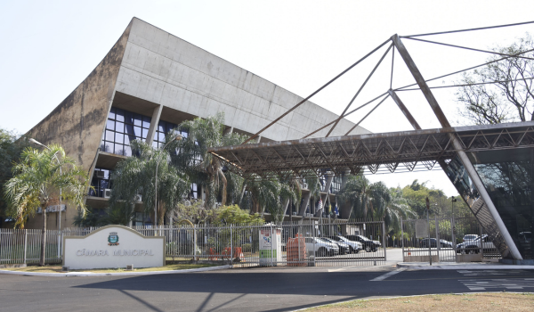 Fachada da Câmara Municipal de Ribeirão Preto, com fachada moderna em concreto, cercada por palmeiras e estrutura metálica sobre a entrada principal.