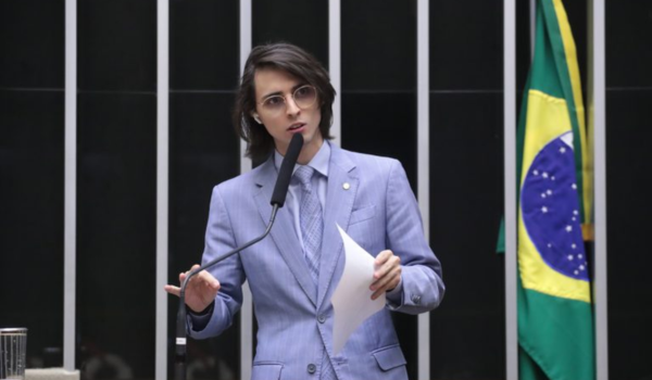Pessoa de terno azul claro falando ao microfone em plenário com a bandeira do Brasil ao fundo.