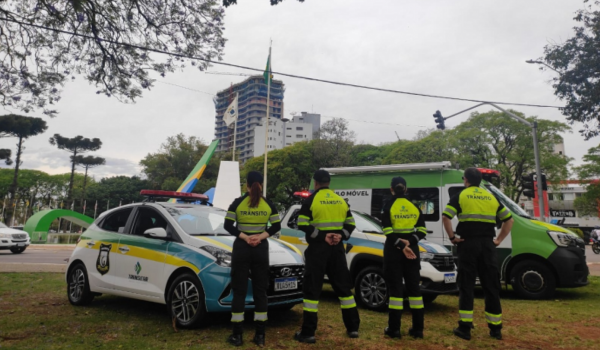 Agentes de trânsito uniformizados em frente a viaturas da Superintendência de Trânsito de Maringá (Setran), posicionados de costas, com bandeiras e prédio em construção ao fundo.