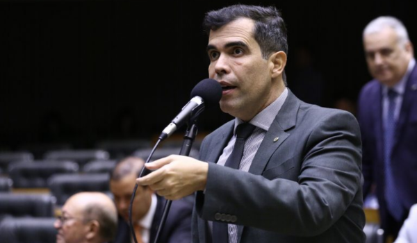 Homem de terno cinza escuro fala ao microfone em um plenário, gesticulando com a mão direita. Outras pessoas estão sentadas ao fundo.