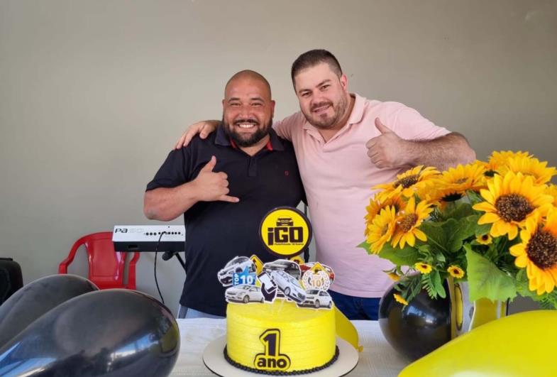 Dois homens sorrindo atrás de um bolo amarelo decorado com carros e a inscrição "1 ano", com balões pretos e flores amarelas ao redor.