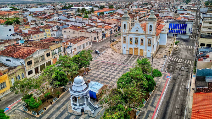 Vista aérea de uma praça central com coreto, árvores, calçamento quadriculado e igreja histórica ao fundo, rodeada por construções urbanas.