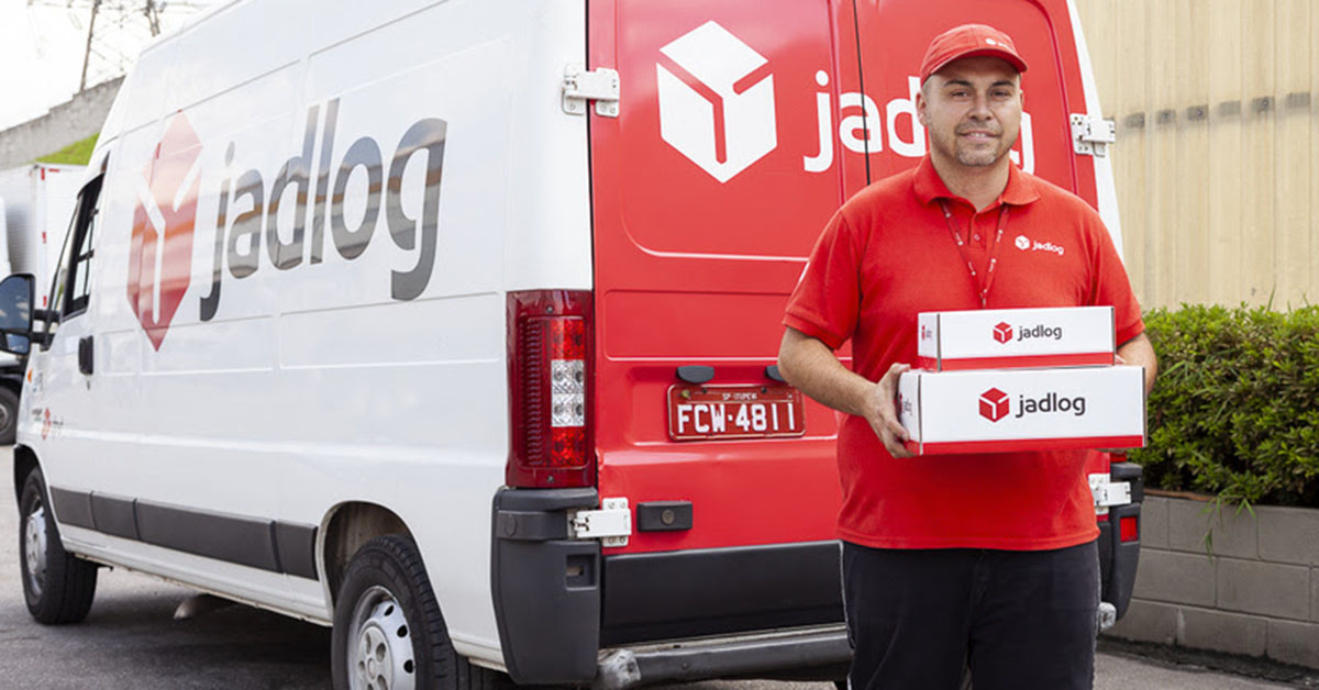 Entregador da Jadlog segurando encomenda em caixa em frente a carro da empresa.