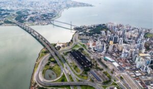 Vista aérea de Florianópolis mostrando a Ponte Pedro Ivo Campos ligando a ilha ao continente, com prédios altos e áreas residenciais próximas ao mar.