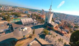 Vista aérea de uma praça central em uma cidade, com prédios altos, uma torre com antena e uma estrutura circular moderna no primeiro plano.