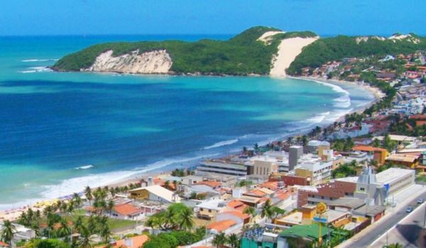 Vista aérea da Praia de Ponta Negra, em Natal, com mar azul esverdeado, faixa de areia clara, edifícios à beira-mar e o Morro do Careca ao fundo.