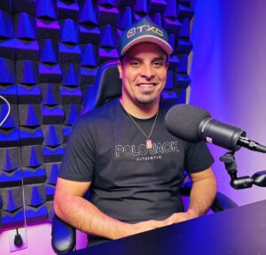 Elvis Santos sentado em uma mesa de podcasting, sorrindo em frente a um microfone.