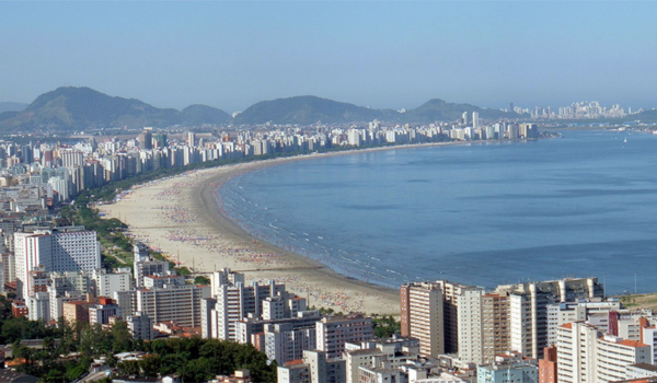Vista panorâmica da orla de Santos, com praia extensa, mar calmo e diversos prédios residenciais e comerciais alinhados à beira-mar.