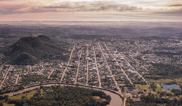 Vista aérea de uma cidade com ruas retas e paralelas, rodeada por áreas verdes, montanhas e um rio sinuoso em primeiro plano.